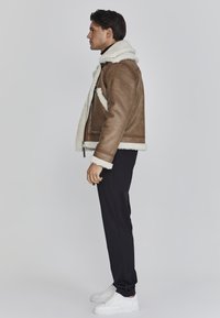 Chaqueta de cuero marrón con forro y puños de piel de oveja blanca, con cremallera frontal y bolsillos laterales, combinada con pantalones negros y zapatillas blancas.