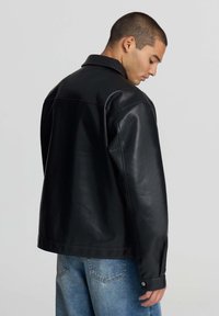 Giovane uomo con capelli corti, che indossa una giacca di pelle nera e jeans blu, che guarda verso il basso su uno sfondo chiaro e semplice.