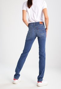 Jeans de mezclilla azul con un corte ajustado, que cuentan con bolsillos traseros, un parche con el logo en la cinturilla, y combinados con zapatillas blancas y una camisa blanca sencilla.