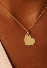 Gouden hartvormige hangerketting op een fijne ketting, voor een bruine achtergrond. De hanger is glad met een reflecterende oppervlakte.