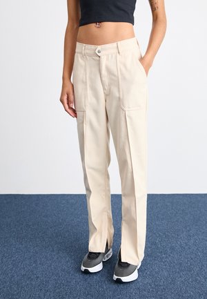 Beige canvas trousers with a straight fit, featuring two front pockets and visible stitching. Paired with black and white athletic shoes.