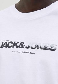 Baltas kokvilnas T-krekls ar apaļu kakla izgriezumu, uz kura ir liels melns un pelēks grafiskais attēls ar uzrakstu "JACK & JONES" un mazāku tekstu zem tā.