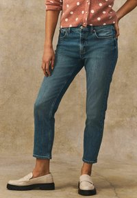 Siniset farkut slim fit -leikkauksella ja lyhyellä pituudella, yhdistettynä kreemivalkoisiin mokkasiineihin. Pehmeä teksturoitu vaaleanpunainen pilkullinen neuletakki päällä.