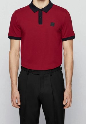 Homme portant un polo rouge avec un col noir et des bordures de manches noires, rentré dans un pantalon noir plissé avec une ceinture noire.