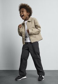 Veste zippée beige sur chemise blanche, pantalons noirs amples et baskets noires. Tissu texturé avec des manches à revers et un détail de logo.