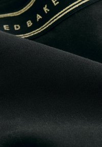 Zwarte stof met een gladde textuur, voorzien van gouden stiksel langs de rand en een label. De stof vertoont een consistente, fijne ribbelpatroon.