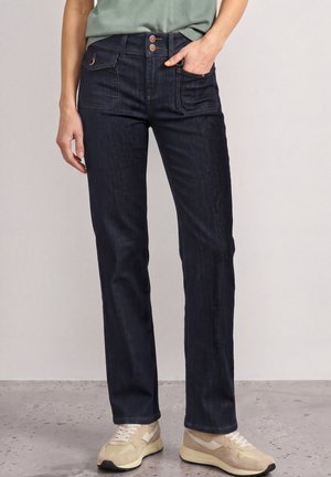 Persona che indossa jeans blu scuro a gamba dritta con tasche frontali con patta e sneaker beige, in piedi su un pavimento grigio chiazzato.