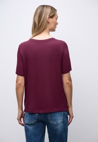 Burgundowa bluzka z krótkim rękawem z miękkiego materiału, z okrągłym dekoltem, luźnym krojem i bocznymi rozcięciami, zestawiona z jasnoniebieskimi dżinsami.