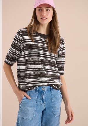 Frau trägt einen braun-weiß gestreiften Kurzarmpullover, hellblaue hoch taillierte Jeans und eine pinke Baseballkappe, steht mit einer Hand in der Tasche.
