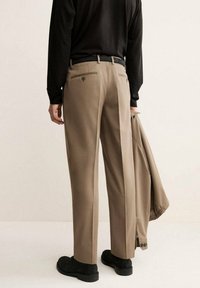 Pantaloni beige su misura con vestibilità dritta, tasche posteriori con dettagli a bottone e una piega che scorre lungo le gambe, indossati con un top nero.