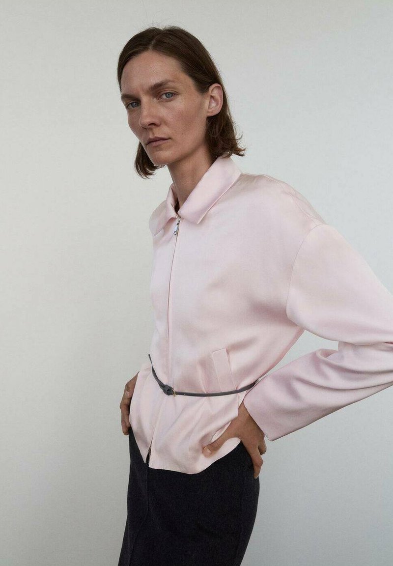 Veste en satin rose clair avec fermeture éclair, col pointu, manches longues et taille cintrée. Dotée de poches latérales et d'une texture lisse.