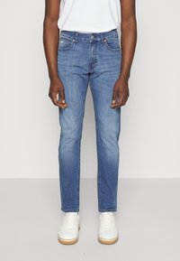 Světlé modré džíny s rovnou nohavicí, pěti kapsami a jemnými švy, nošené s bílými teniskami.