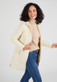 Abrigo acolchado de un beige claro con capucha, cierre de cremallera, bolsillos laterales y tejido texturizado, usado sobre un cuello de tortuga beige y unos vaqueros azules.