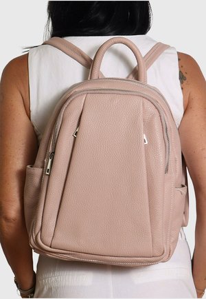 Femme portant un sac à dos en cuir rose blush avec des fermetures éclair argentées, vue de dos sur un fond uni.