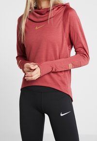 Maroon long-sleeve athletic top with a hood, gold logo, and thumbholes, paired with black leggings featuring a white logo. Smooth texture.