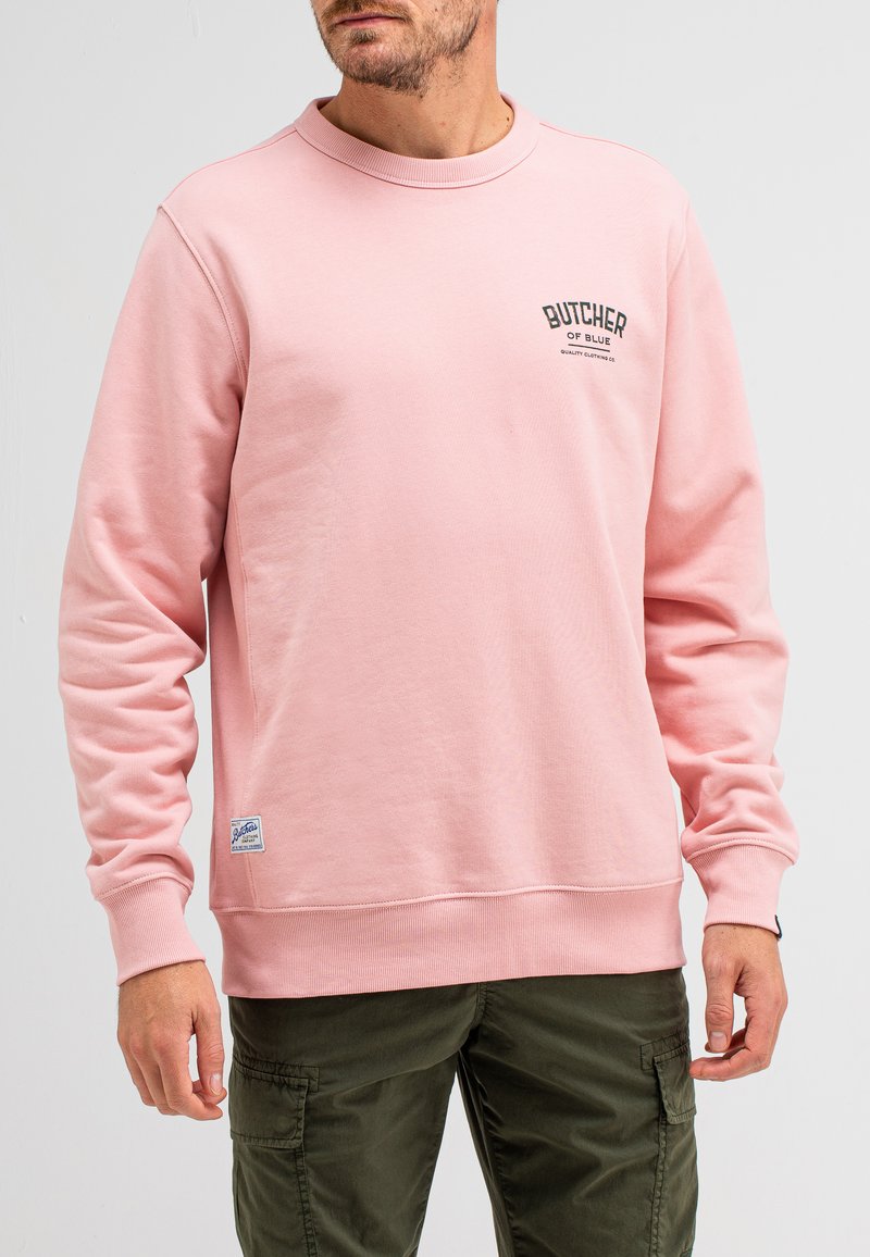 Lichtroze crewneck sweatshirt van een katoenmix, met een klein zwart logo op de borst en geribbelde manchetten. Olijfgroene broek wordt gedragen.