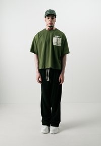 Olivenfarget overdimensjonert t-skjorte med brystlomme, sammen med svarte joggebukser og hvite klumpete sneakers. Tilbehøret inkluderer en mørkegrønn caps.