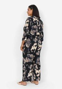 Black jumpsuit with a bold, abstract beige and gray pattern, featuring wide legs and three-quarter sleeves, paired with light flat shoes.
