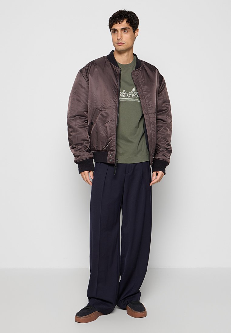 Joven con una chaqueta bomber marrón oscuro, camiseta gráfica verde oliva, pantalones anchos azul marino y zapatillas negras con suelas marrones, de pie