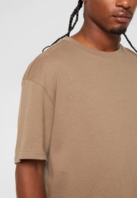 Tricou din bumbac de culoare maro deschis, cu guler rotund și mâneci scurte, având o croială lejeră și o textură moale, surprins într-o imagine detaliată.