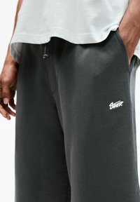 Gray cotton sweatpants with a smooth texture, elastic waistband, and a white logo on the left thigh. Loose-fit design, side pockets visible.