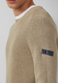 Beige gestrickter Pullover mit strukturiertem Muster, geripptem Kragen und einem schwarzen Etikett am linken Arm, das die Marke und das Gründungsjahr anzeigt.