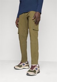 Pantalones cargo en verde oliva con bolsillos laterales, corte recto y detalles de costura visibles, combinados con zapatillas en tonos marrón y verde con un acento púrpura.