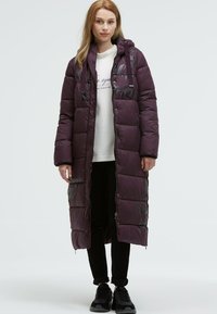 Long, purple puffer coat with zipper and hood, featuring a geometric pattern and front pockets. Worn over a light-colored sweatshirt and black pants.