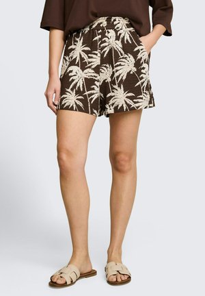 Braune Shorts mit beigefarbenem Palmenmuster, getragen mit beigen Slip-On-Sandalen und einem braunen Oberteil, Hände in den Taschen, stehend vor hellem Hintergrund.