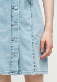 Χέρι που ακουμπάει δίπλα σε φούστα από ανοιχτό μπλε τζιν με πέντε ασημένια κουμπιά κατά μήκος του μπροστινού κέντρου.