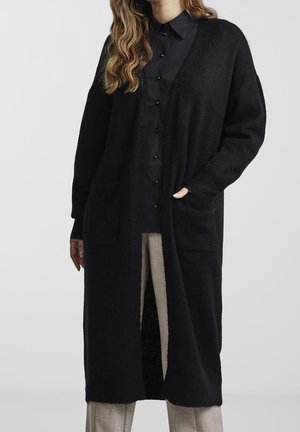 Lång svart cardigan i texturerat tyg med öppen fram, två framfickor och avslappnad passform. Stylad över en skjorta med knappar och byxor.