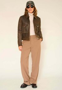 Veste en cuir marron avec deux poches avant, col roulé beige, pantalon marron ample, lunettes de soleil noires et une casquette avec du texte. Textures lisses.