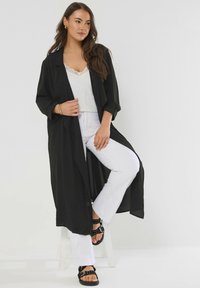 Donna seduta su uno sgabello bianco, indossa un lungo cappotto nero, un top in pizzo bianco, pantaloni bianchi, sandali neri con borchie e orecchini a cerchio dorati.