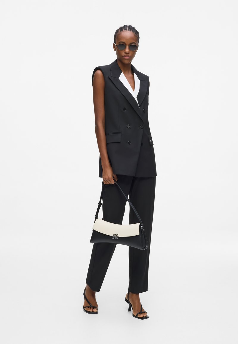 Femme portant un blazer sans manches noir et un pantalon, tenant un sac à main noir et blanc, avec des lunettes de soleil et des sandales noires à talons.