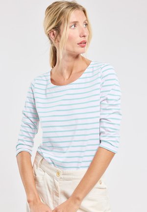 Femme blonde regardant à droite, portant un haut blanc à manches longues avec de fines rayures horizontales bleu clair et un pantalon beige clair avec un bouton.