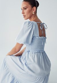 Svetlo modra obleka s napihnjenimi rokavi in smučasto zgornjim delom, ki ima detajl v obliki mašnje na hrbtni strani ter večplastno krilo. Gladka tekstura tkanine.