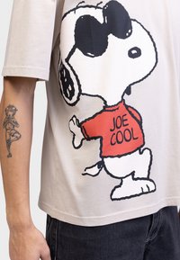 Bež lühikeste varrukatega T-särk, millel on suur musta kontuuriga joonistus Snoopy'st punases "JOE COOL" särgis; puuvillane materjal, mugav lõige.