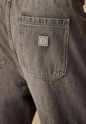Jean en denim gris avec une poche arrière en patch portant une étiquette blanche portant l'inscription « SOLSTICE ». Le tissu présente une légère texture et un aspect usé.