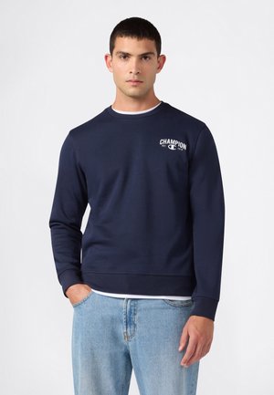 Uomo che indossa una felpa blu navy con il logo "Champion" sul petto e jeans azzurri, in piedi davanti a uno sfondo bianco uniforme.