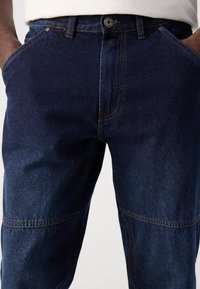 Dark blue denim jeans featuring contrast stitching, a button closure, and five-pocket design, with a slightly worn texture.