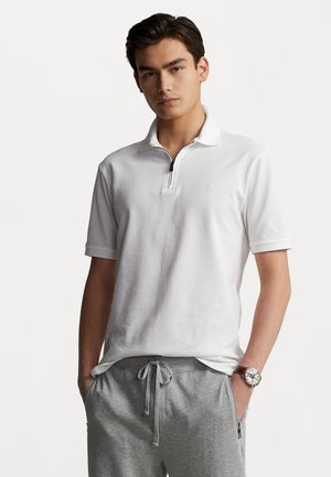 Camisa polo blanca con cuello con cremallera, material de algodón, mangas cortas y un pequeño logo. Combinada con pantalones de chándal grises y un reloj.