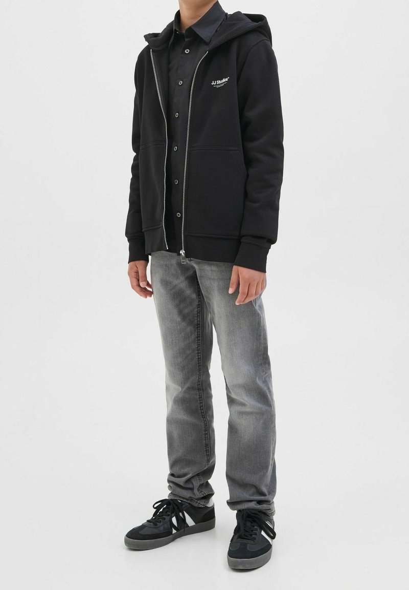 Garçon portant un sweat à capuche noir zippé, une chemise noire à boutons, un jean gris et des baskets noires, debout sur un fond blanc.