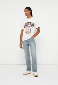 Bela bombažna majica s napisom "Harvard University" v maroon barvi, v kombinaciji s svetlo modrimi ravnimi jeans hlačami in bež tenisicami.