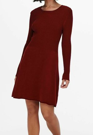 Femme portant une robe rouge foncé côtelée à manches longues et longueur genou, debout devant un fond blanc.