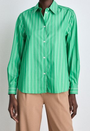 Zielona koszula na guziki z długim rękawem w białe pionowe paski, noszona z beżowymi spodniami, widoczna od połowy klatki piersiowej do ud.