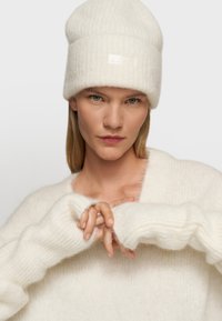 Un bonnet en tricot blanc avec un motif texturé et une étiquette, accompagné d'un doux pull blanc avec des poignets côtelés. Le modèle a les mains croisées.