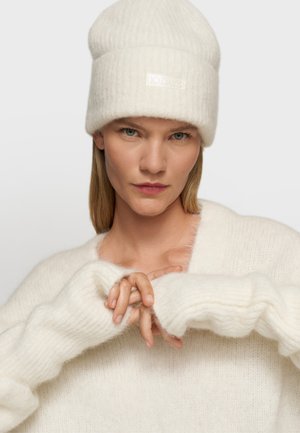 Witte gebreide beanie met een textuurpatroon en label, gecombineerd met een zachte witte trui met ribgebreide manchetten. Model heeft de handen samengevouwen.