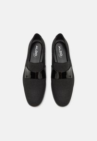 Zapatos negros de tipo slip-on con un upper de tela texturizada, que presentan una banda de acento negra brillante en la parte media del pie y una plantilla acolchada.