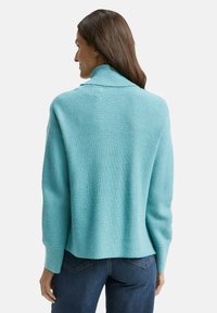 Türkisfarbener gestrickter Pullover mit geripptem Muster, umgeschlagenem Kragen und lockerer Passform, kombiniert mit blauen Jeans.