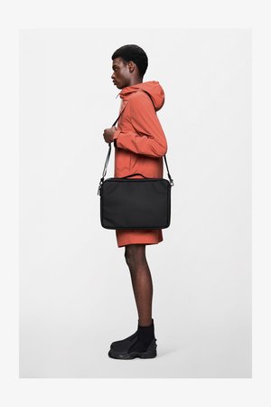 Črno torbo za čez ramo z zgornjim ročajem in nastavljivim trakom. Model nosi svetlo oranžno lahko jakno in črne gležnjarje.